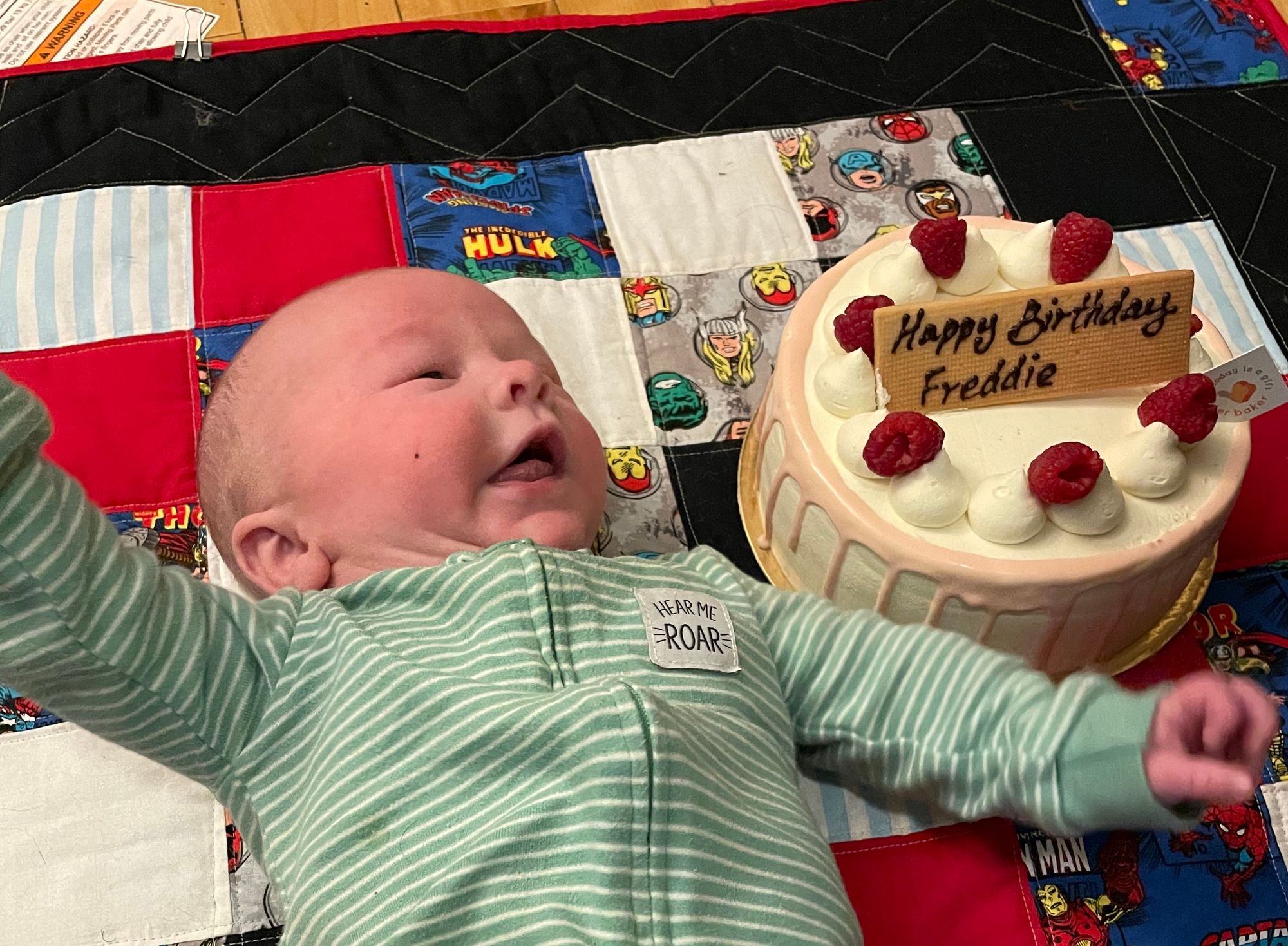 Freddie in a green onesie, with a strange facial expression on his face, next to a cake.